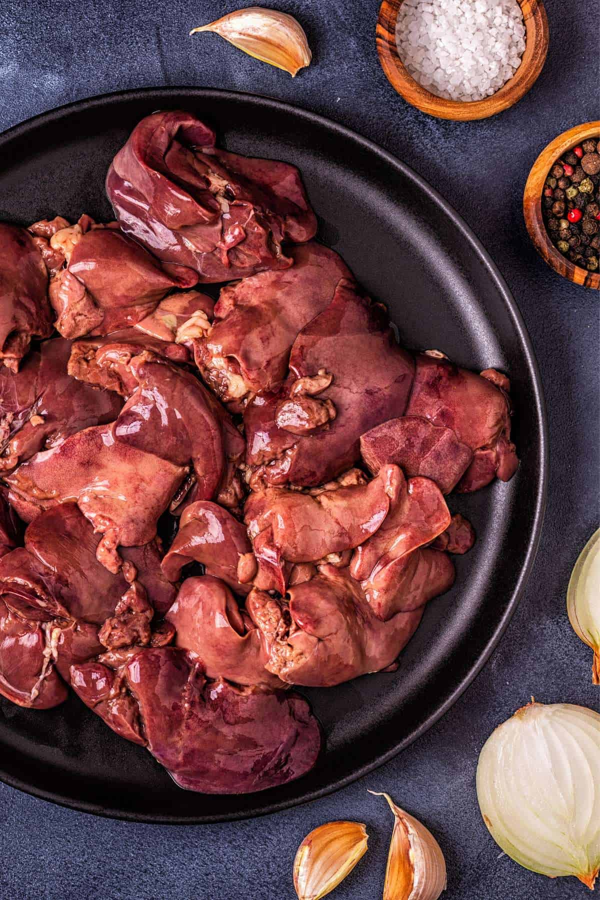 Uncooked Chicken liver on a black plate with garlic, onions, salt and pepper near the plate 